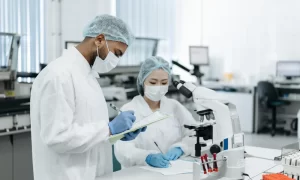 Unos personas con ropa y equipo de laboratorio revisando un documento frente a una mesa con un microscopio
