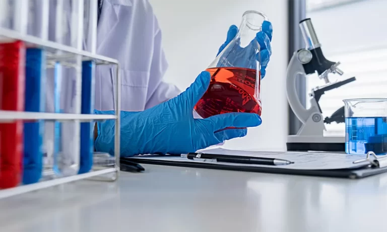 Una mano con guante azul, sosteniendo un matraz con liquido rojo, y un microscopio al fondo