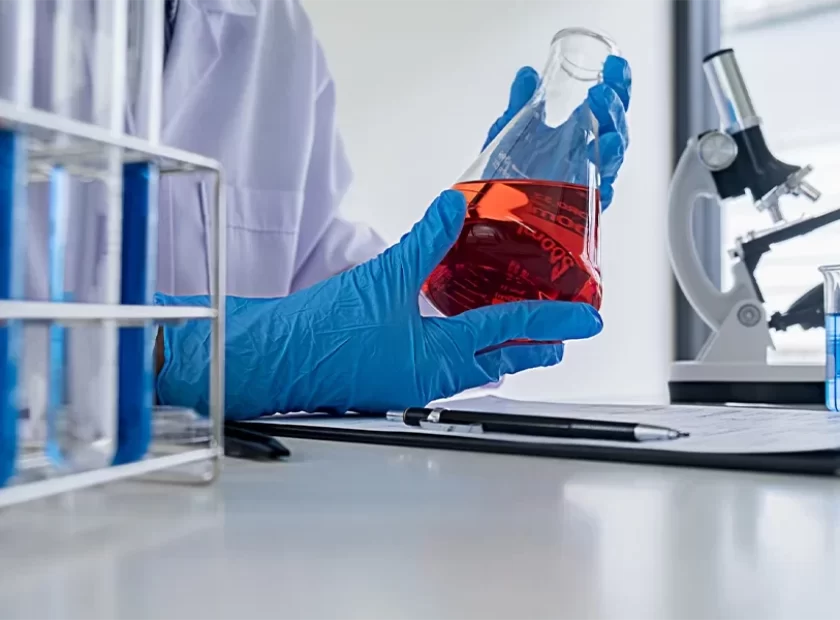 Una mano con guante azul, sosteniendo un matraz con liquido rojo, y un microscopio al fondo